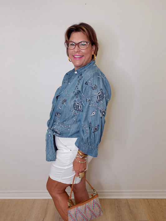 Woman wearing a blue floral shirt and white shorts, standing against a plain wall.