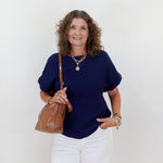 Woman wearing a navy blue top and white pants, holding a brown leather bag against a white background