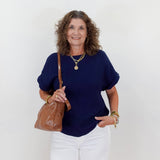 Woman wearing a navy blue top and white pants, holding a brown leather bag against a white background