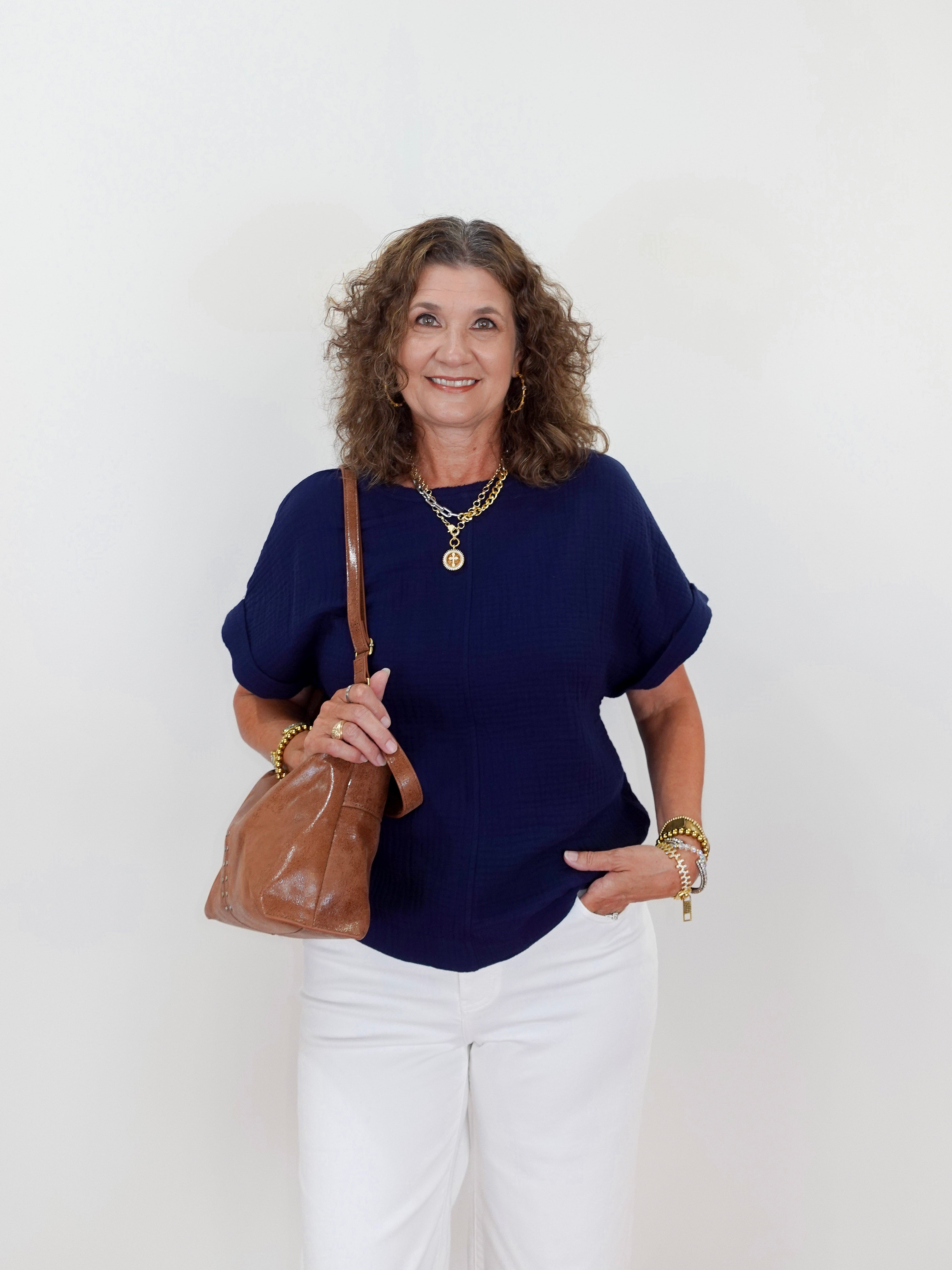 Woman wearing a navy blue top and white pants, holding a brown leather bag against a white background