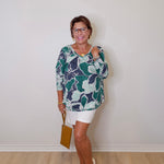 Woman wearing a patterned top and white shorts against a plain wall.