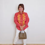 Woman wearing a pink floral blouse, white pants, and holding a handbag against a white wall.
