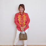 Woman wearing a pink floral blouse, white pants, and holding a handbag against a white wall.