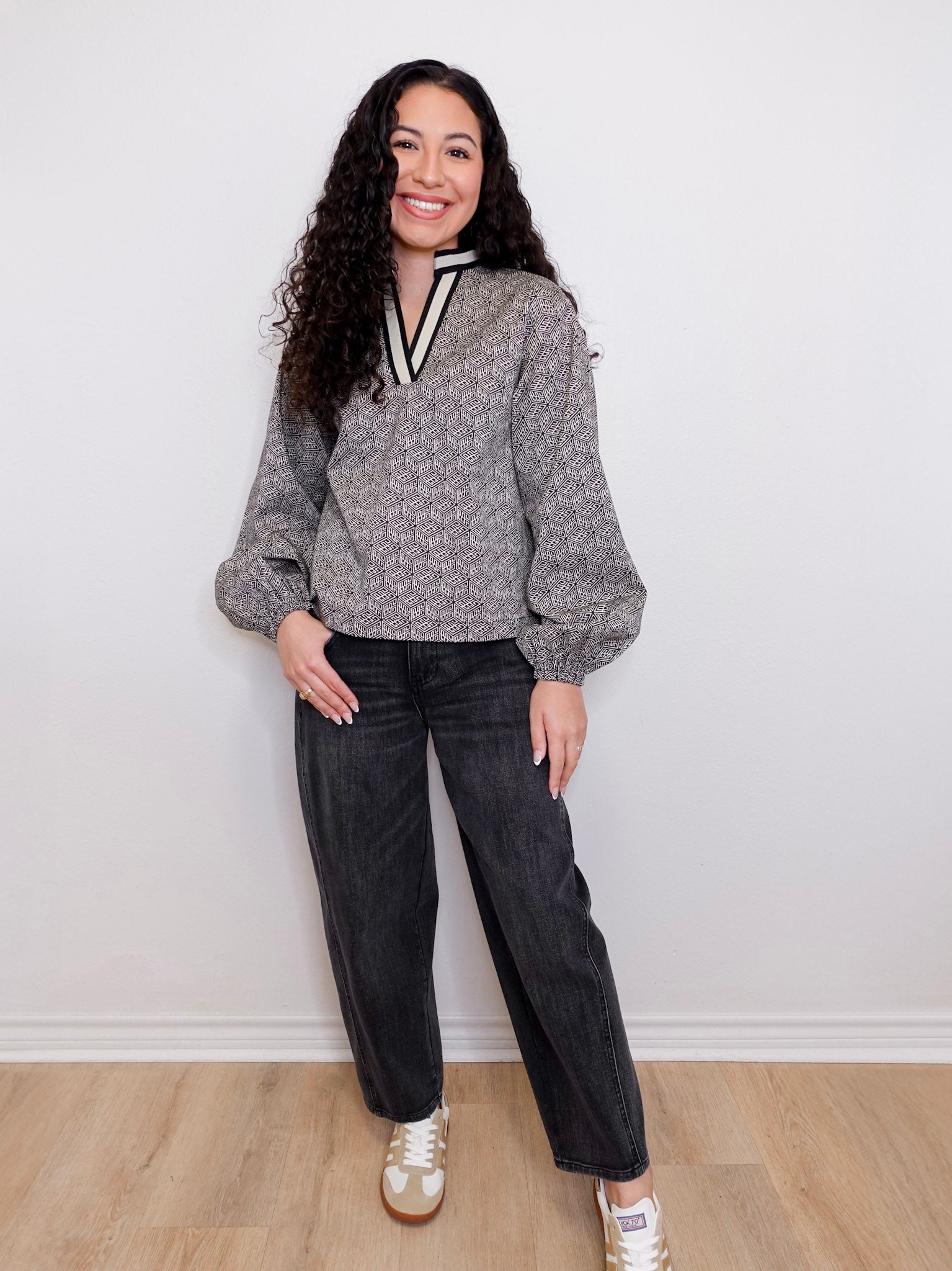Woman wearing a patterned top and black pants against a white wall