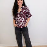 Woman wearing a patterned shirt and black pants against a white wall