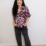 Woman wearing a patterned shirt and black pants against a white wall