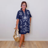 Woman wearing a navy dress with white floral patterns, holding a gold handbag against a plain background.