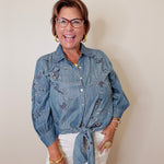 Woman wearing a blue denim shirt with floral embroidery and white shorts against a plain background
