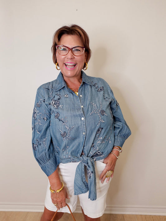 Woman wearing a blue denim shirt with floral embroidery and white shorts against a plain background