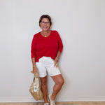 Woman in a red top and white shorts posing against a plain wall.
