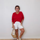 Woman in a red top and white shorts posing against a plain wall.