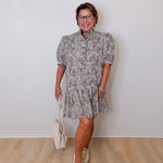 Woman wearing a patterned dress with white sneakers and a white bag against a plain wall.