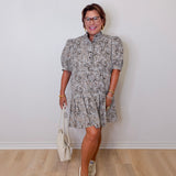 Woman wearing a patterned dress with white sneakers and a white bag against a plain wall.