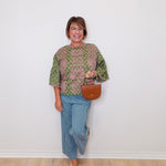 Woman wearing a green patterned top and blue jeans, holding a brown bag against a white wall.