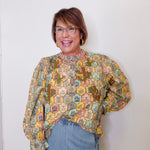 Woman wearing a patterned blouse and jeans against a white wall
