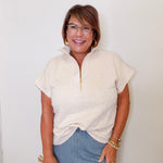 Woman wearing a beige short-sleeve shirt and blue jeans against a white wall.
