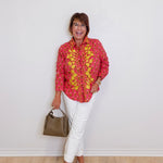 Woman wearing a pink floral blouse and white pants, standing against a plain white wall.