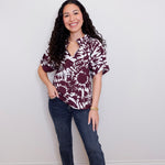 Woman wearing a patterned blouse and jeans standing against a plain wall.