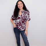 Woman wearing a patterned blouse and jeans standing against a plain wall.
