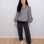 Woman wearing a gray sweater with a V-neck and dark jeans, standing against a white wall.