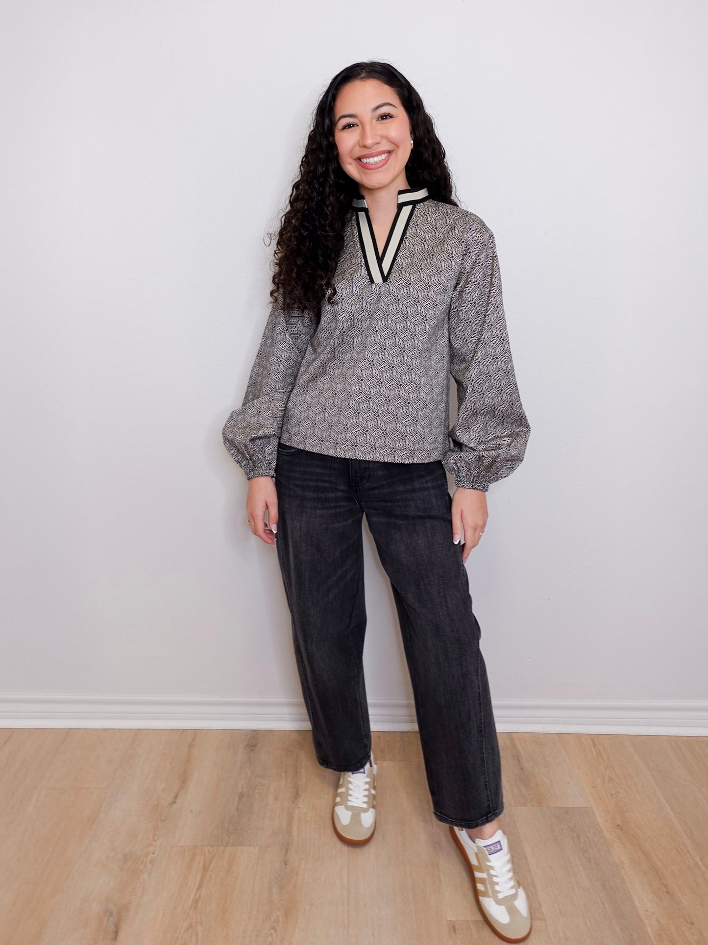 Woman wearing a gray sweater with a V-neck and dark jeans, standing against a white wall.