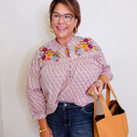 Woman wearing a patterned blouse with floral embroidery, holding a tan handbag against a white background