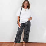 Woman wearing a white top and black patterned pants standing against a white wall.