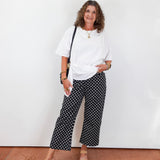 Woman wearing a white top and black patterned pants standing against a white wall.