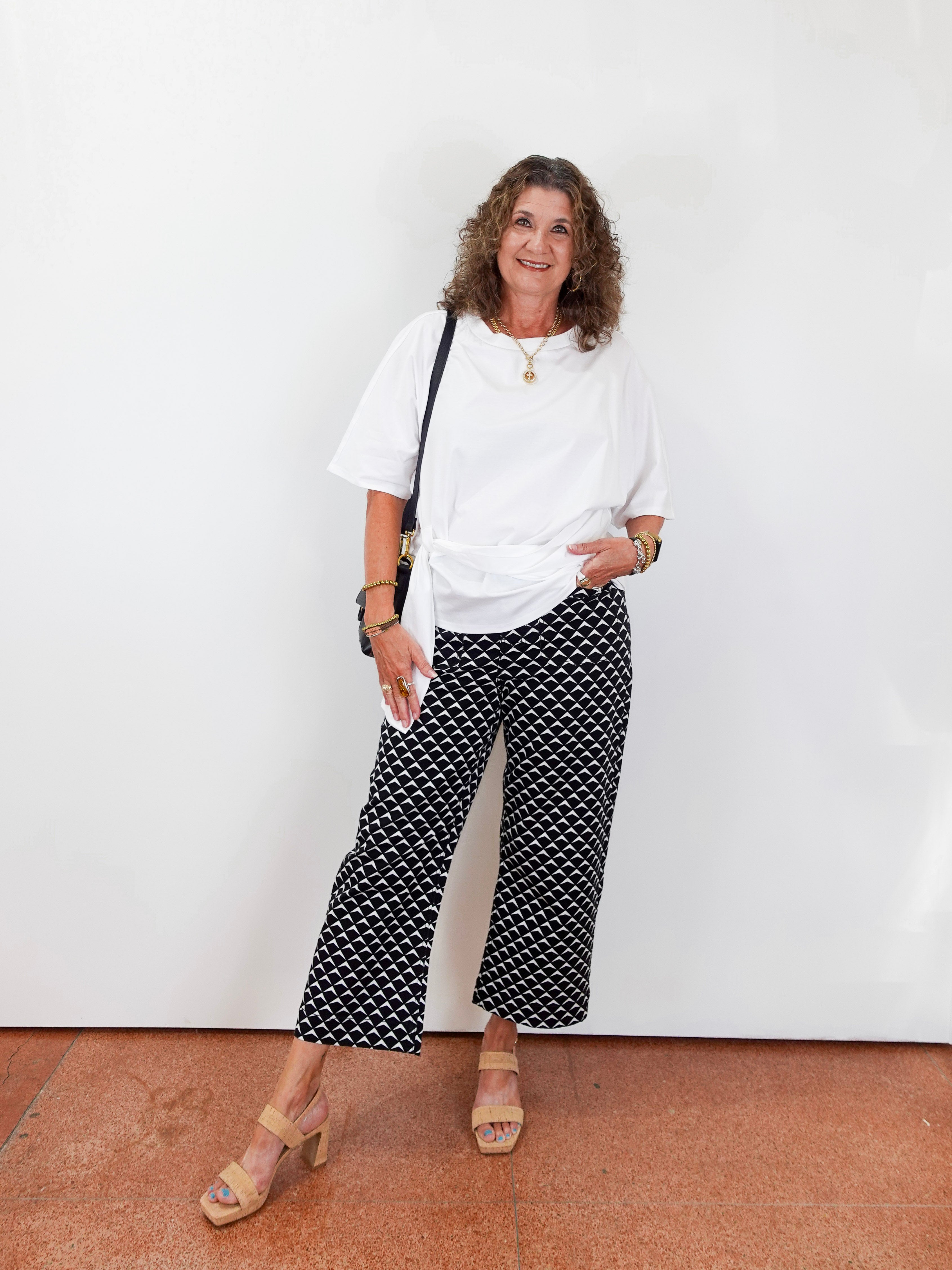 Woman wearing a white top and black patterned pants standing against a white wall.