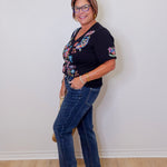 Woman wearing a black floral top and blue jeans against a white wall.