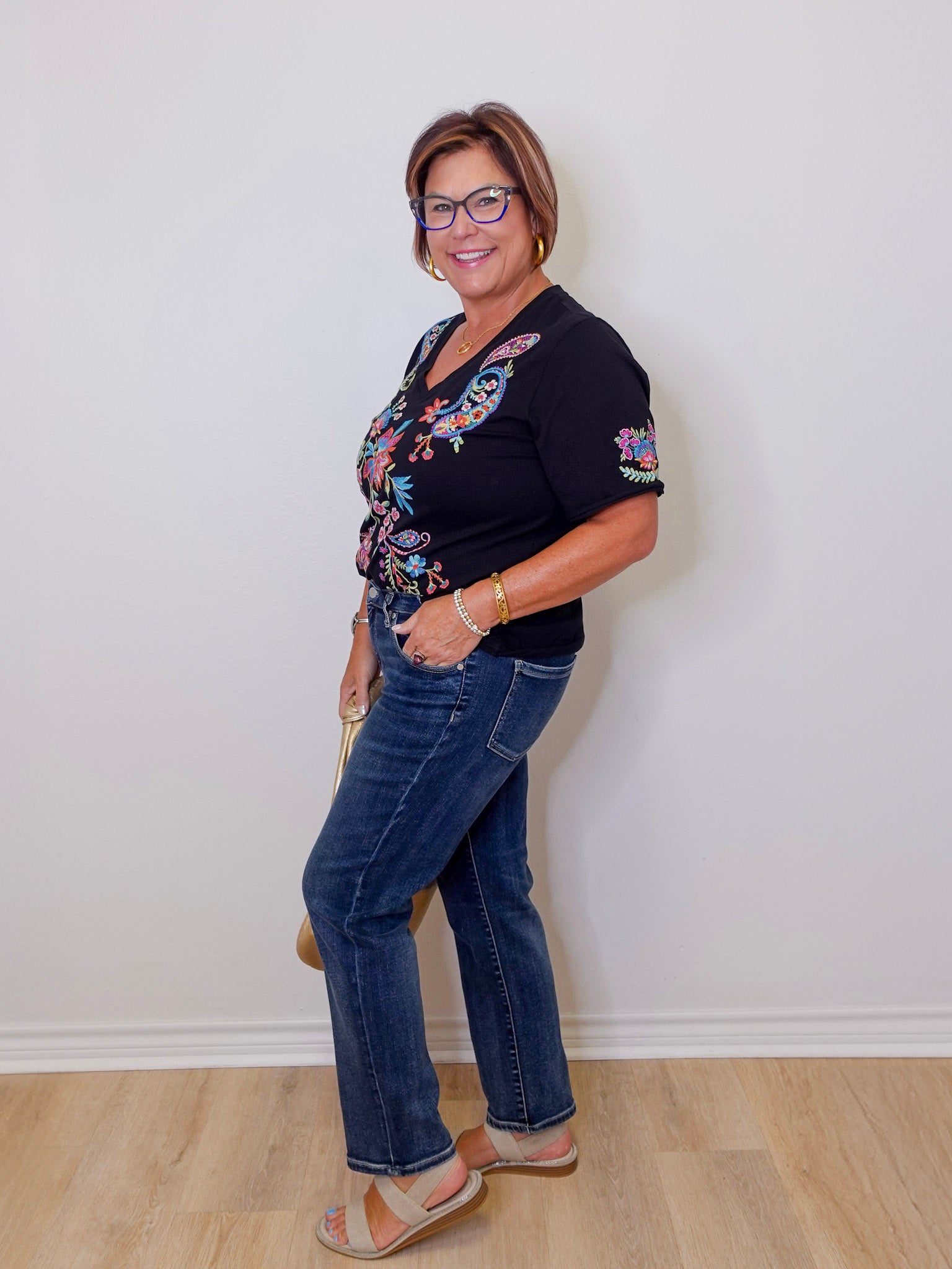Woman wearing a black floral top and blue jeans against a white wall.