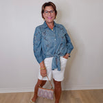 Woman wearing a denim jacket and white shorts standing against a plain wall.