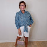Woman wearing a denim jacket and white shorts standing against a plain wall.