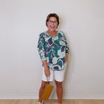 Woman wearing a patterned top and white shorts against a plain wall.