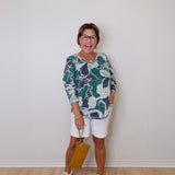 Woman wearing a patterned top and white shorts against a plain wall.