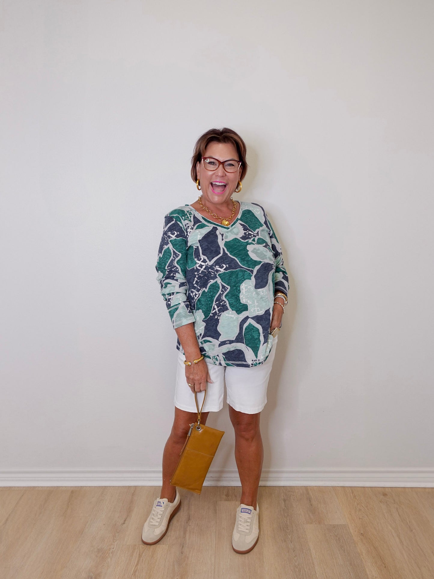 Woman wearing a patterned top and white shorts against a plain wall.