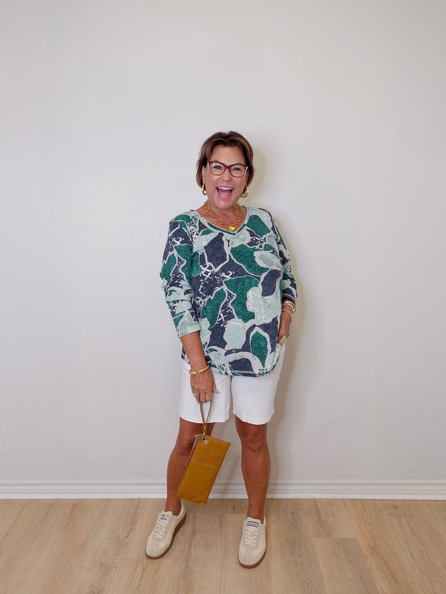 Woman wearing a patterned top and white shorts against a plain wall.