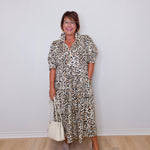 Woman wearing a leopard print dress holding a white handbag against a plain background