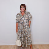 Woman wearing a leopard print dress holding a white handbag against a plain background