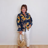 Woman wearing a patterned shirt and white pants, holding a gold handbag against a plain background