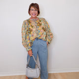 Woman wearing a floral blouse and blue jeans, holding a gray handbag against a white wall.