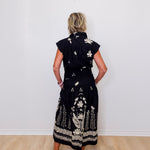 Woman wearing a black dress with white floral patterns against a white wall.
