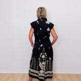 Woman wearing a black dress with white floral patterns against a white wall.