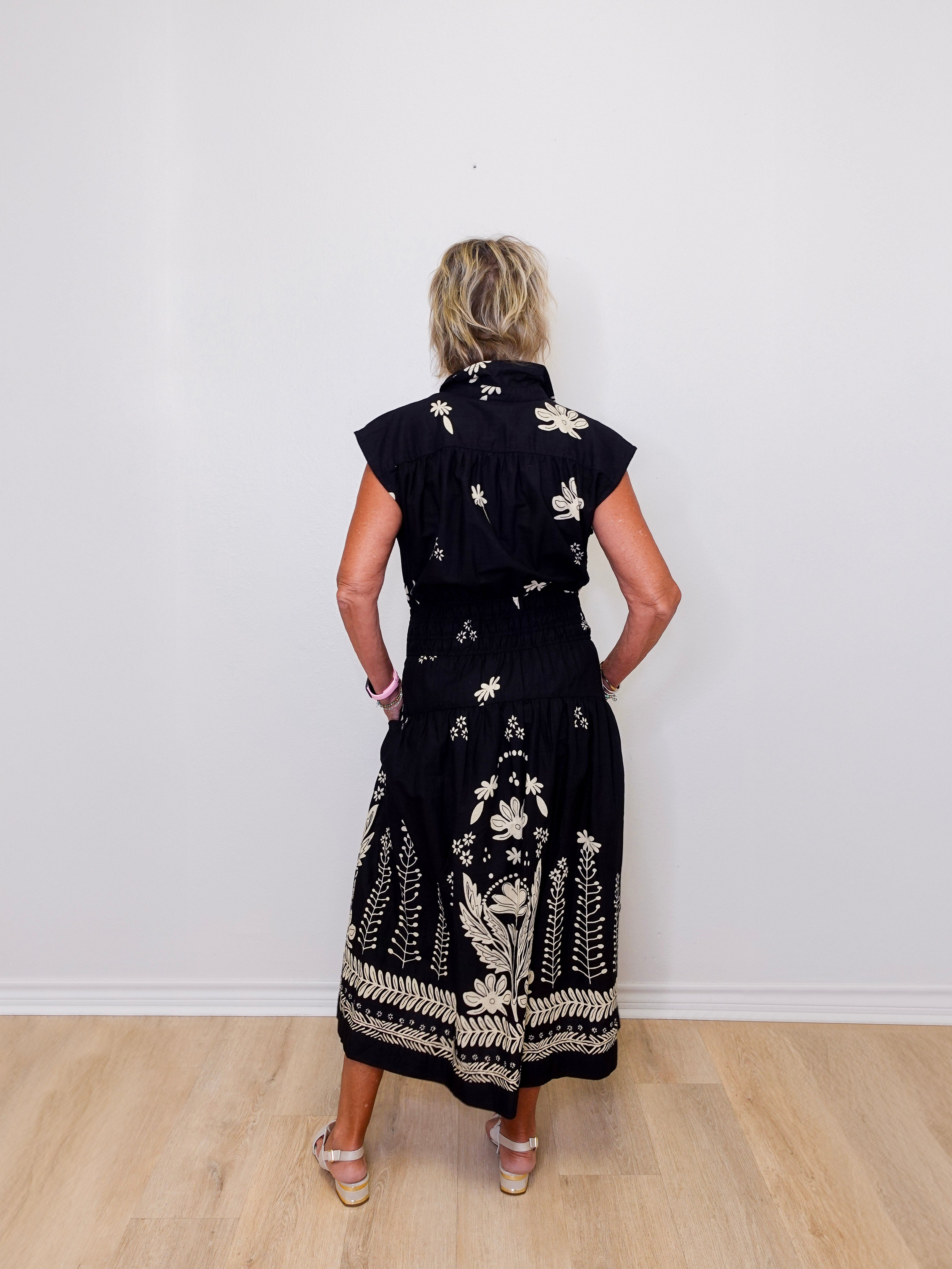 Woman wearing a black dress with white floral patterns against a white wall.