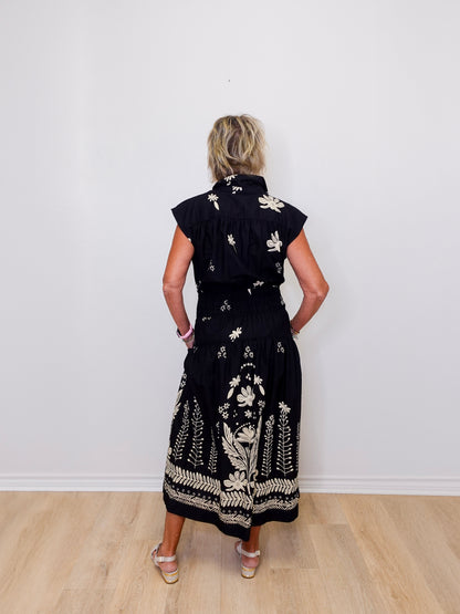 Woman wearing a black dress with white floral patterns against a white wall.