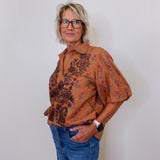 Woman wearing a patterned brown shirt and blue jeans against a white wall