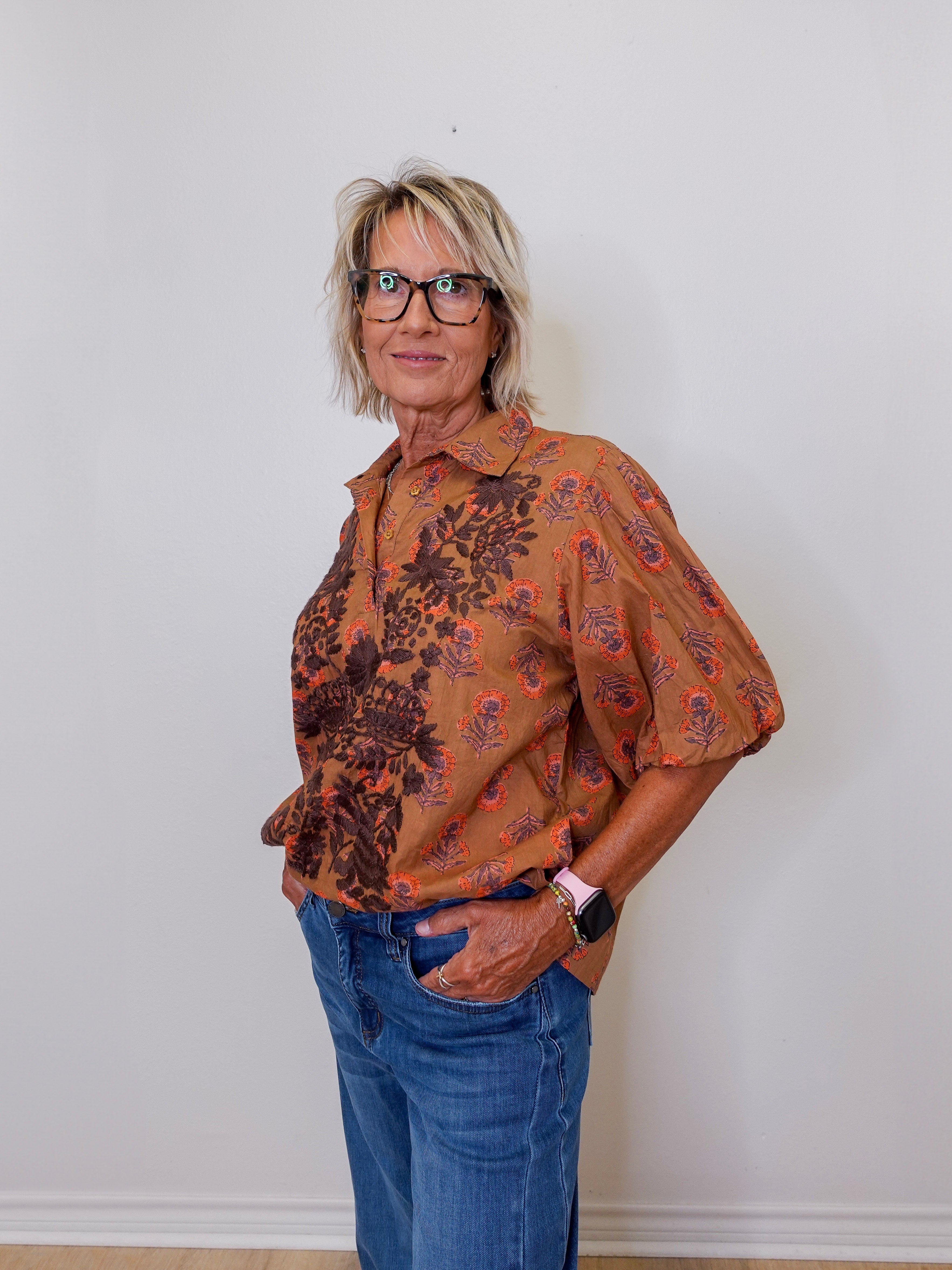 Woman wearing a patterned brown shirt and blue jeans against a white wall