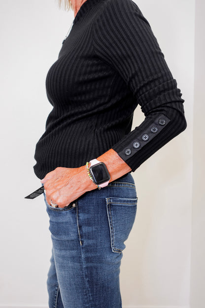 Person wearing a black ribbed sweater with button details on the sleeves, paired with blue jeans.