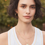 Woman wearing a white tank top with a logo, layered necklaces, and hoop earrings against a blurred natural background.
