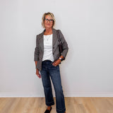 Woman standing against a white wall wearing a brown blazer, white shirt, blue jeans, and brown shoes.